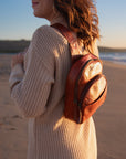 A woman on a sandy beach at sunset wearing a small brown leather backpack, shown from the side and back. The backpack has a smooth brown leather body with a lighter beige canvas front pocket.