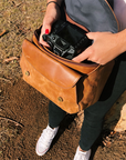 Person placing a camera into a brown leather bag outdoors.