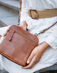 Brown leather handbag held by a person wearing a white dress with a brown belt.