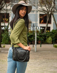 Woman wearing a green shirt, blue jeans, and a wide-brimmed hat, holding a black handbag on a street.
