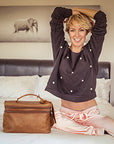 Woman sitting on a bed with a brown bag next to her, wearing a dark sweater and light skirt.