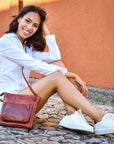 Woman sitting on a stone bench with a brown leather bag, wearing a white shirt and white sneakers.