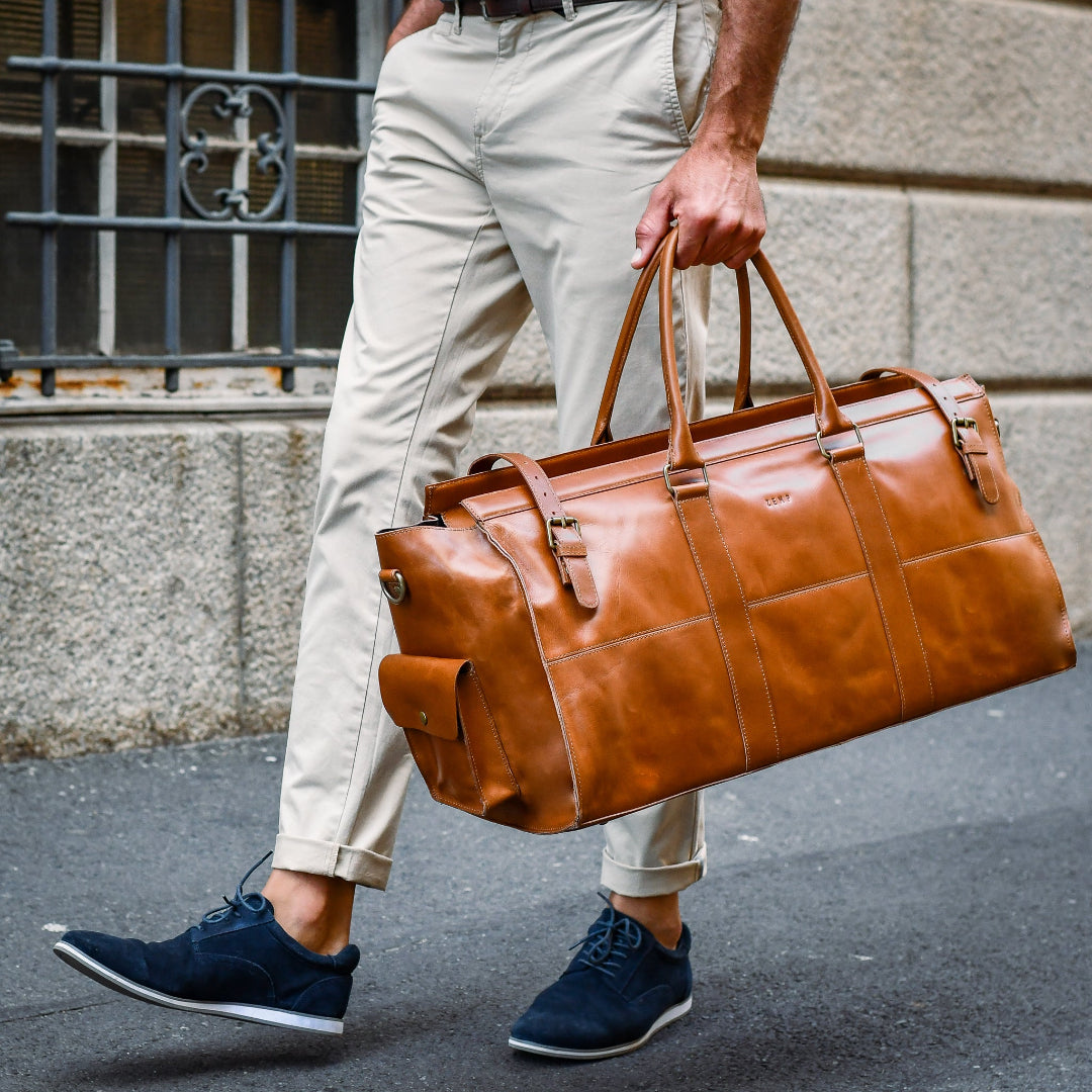 Person walking outdoors holding a brown leather duffel bag.