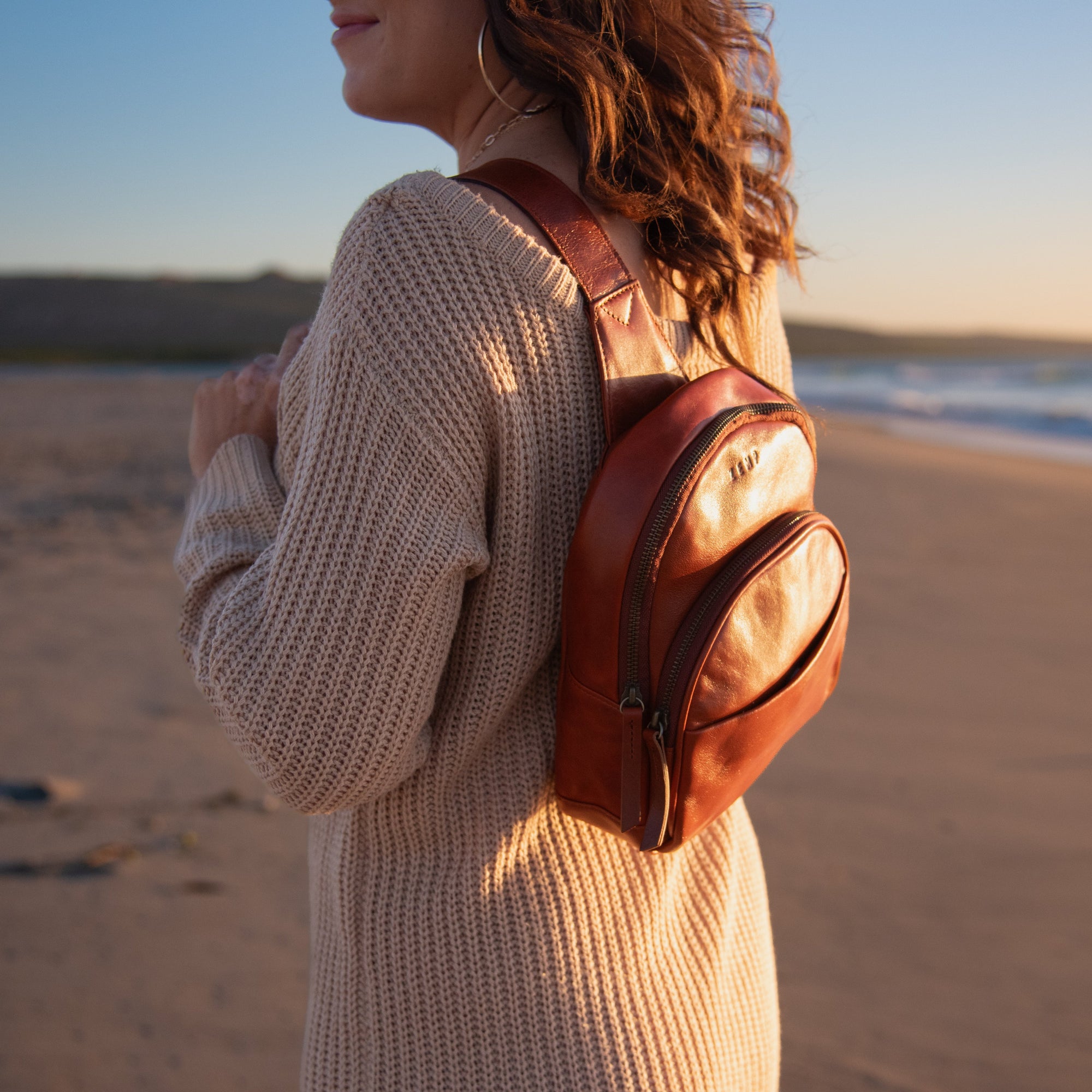 A woman on a sandy beach at sunset wearing a small brown leather backpack, shown from the side and back. The backpack has a smooth brown leather body with a lighter beige canvas front pocket.
