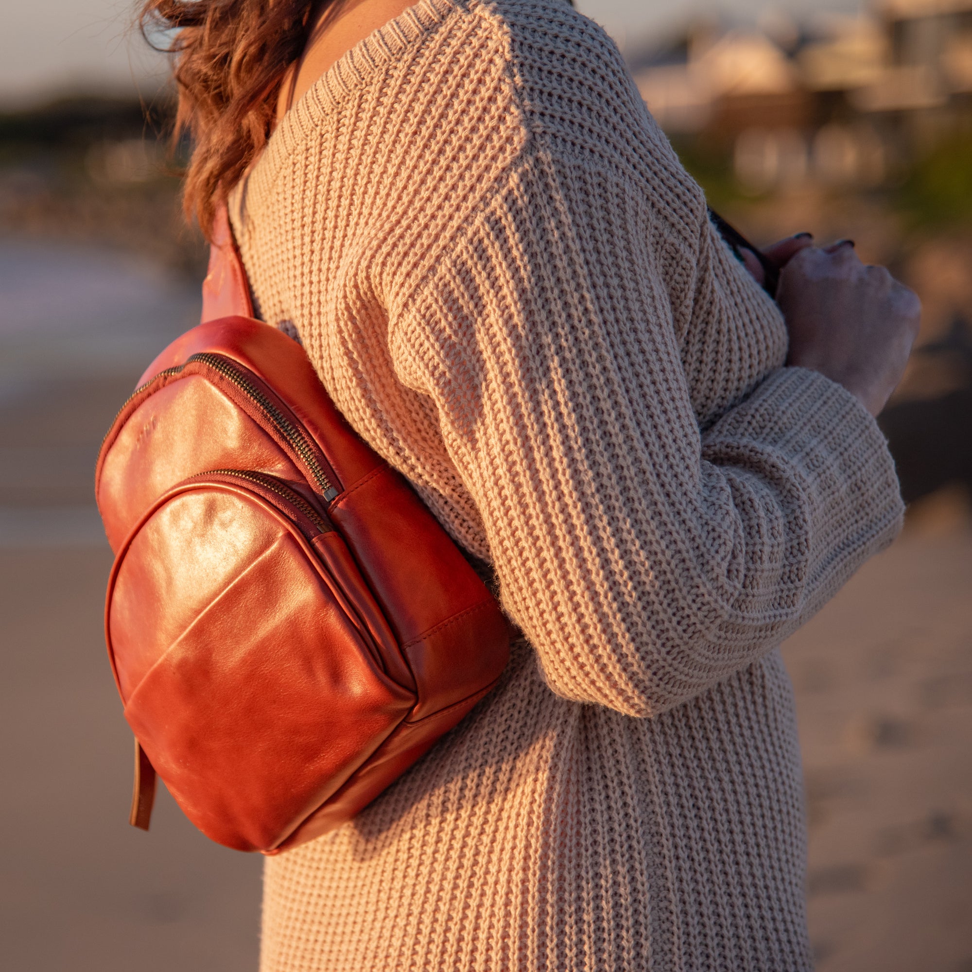 A person wearing a beige ribbed knit sweater carries a small reddish-brown leather backpack on their shoulder in an outdoor setting at sunset.