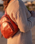 A person wearing a beige ribbed knit sweater carries a small reddish-brown leather backpack on their shoulder in an outdoor setting at sunset.