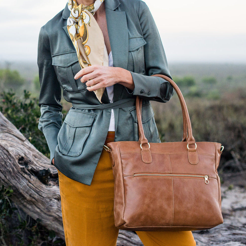 Person holding a brown leather handbag in a natural setting with a hat and scarf.
