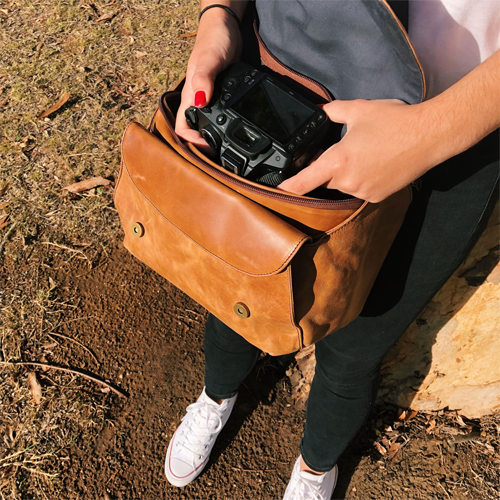 Person placing a camera into a brown leather bag outdoors.