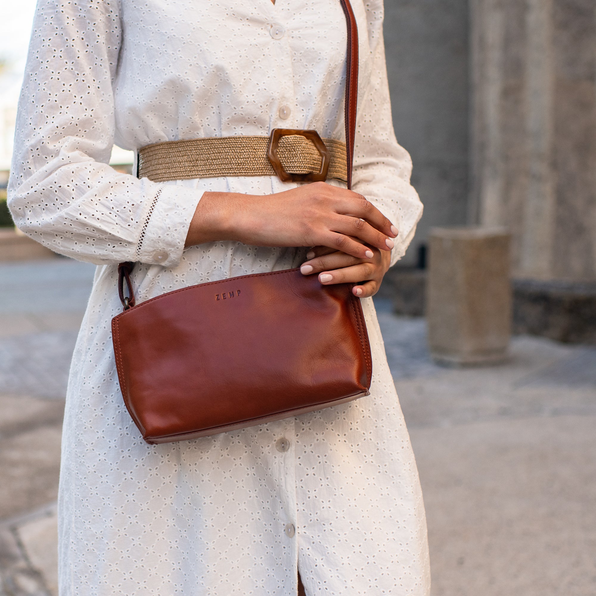 A person wearing a white eyelet dress and a woven belt carries a brown leather crossbody bag with a small embossed logo, photographed outdoors against a blurred stone background.