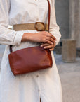 A person wearing a white eyelet dress and a woven belt carries a brown leather crossbody bag with a small embossed logo, photographed outdoors against a blurred stone background.