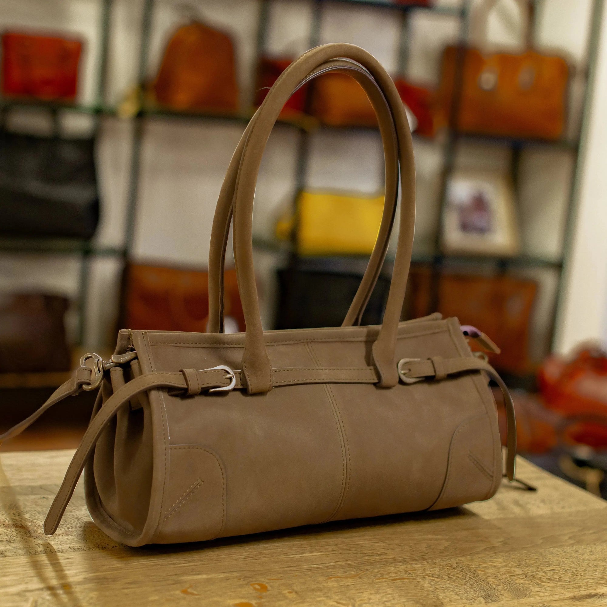 Brown leather handbag on a wooden surface with a blurred background of other bags.