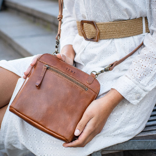 Brown leather handbag held by a person wearing a white dress with a brown belt.