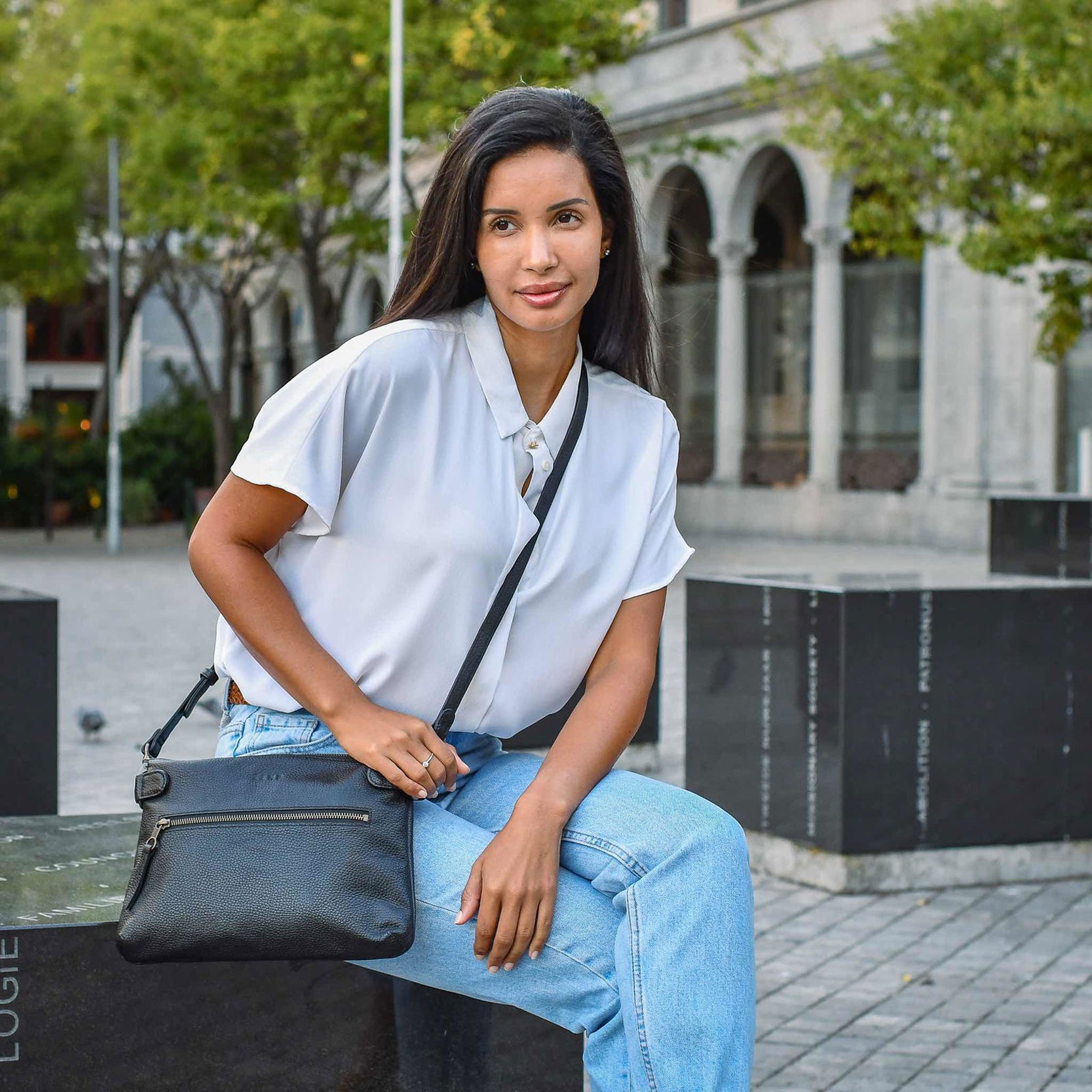 Woman sitting outdoors holding a black handbag, wearing a white shirt and blue jeans.