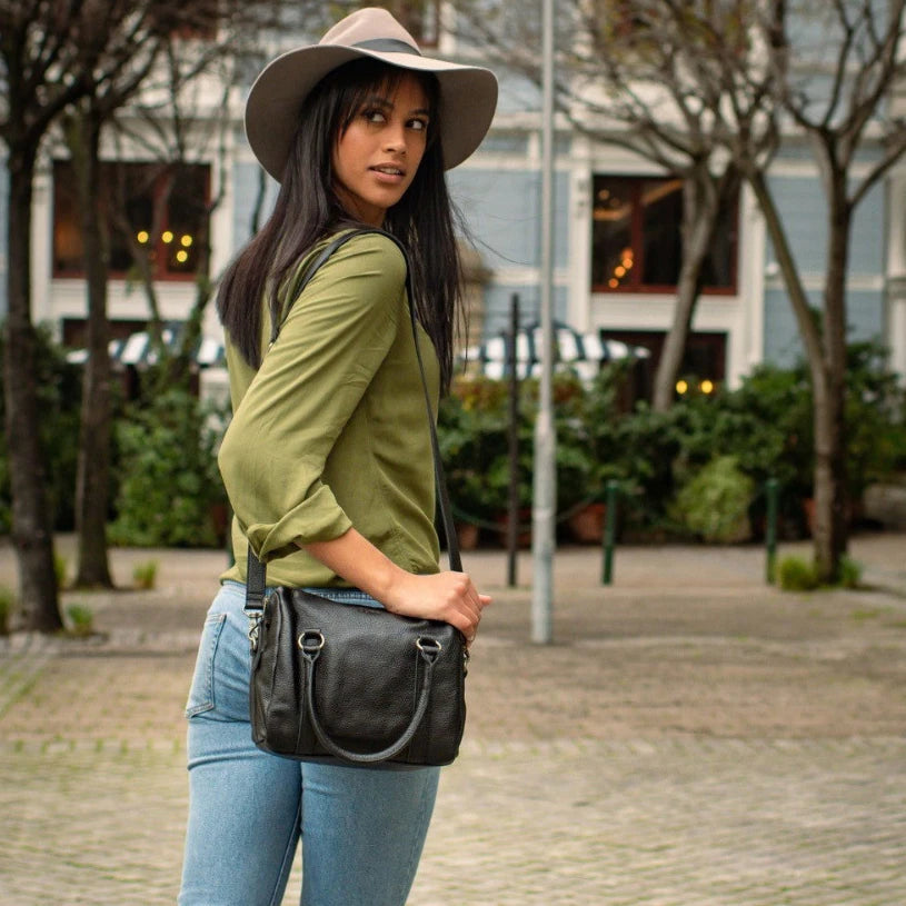 Woman wearing a green shirt, blue jeans, and a wide-brimmed hat, holding a black handbag on a street.