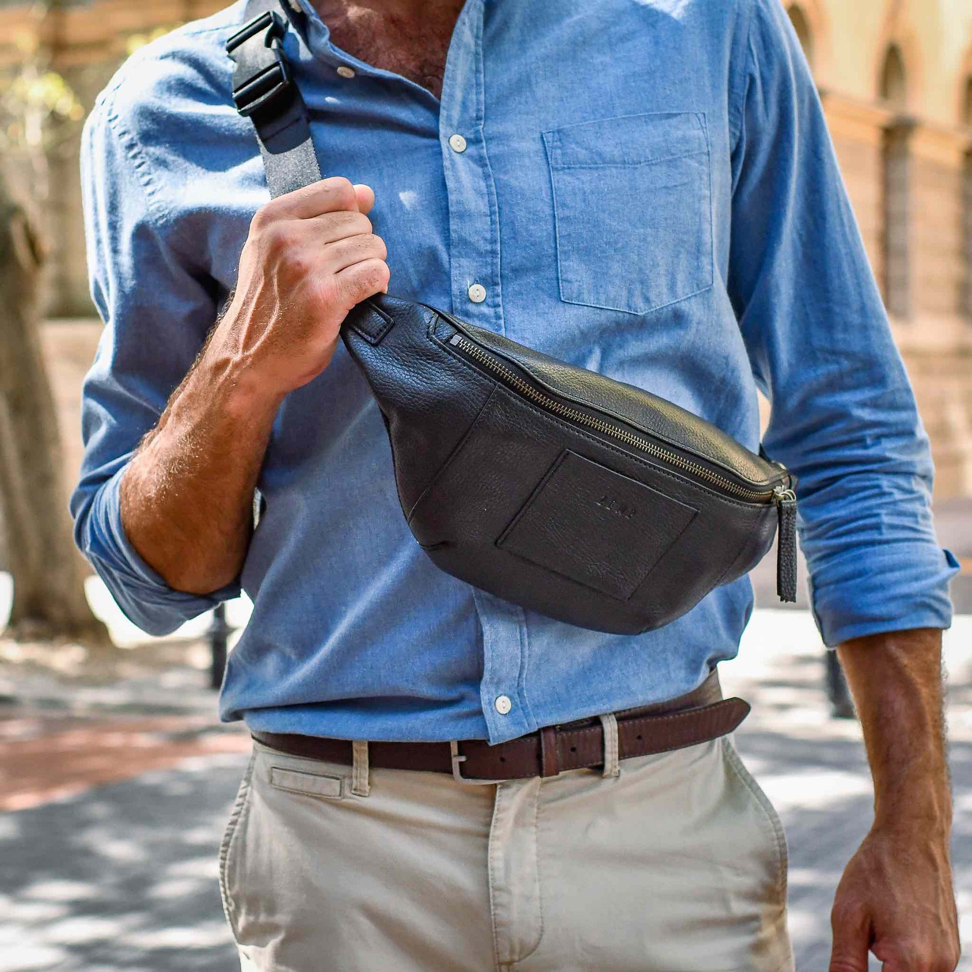 Person wearing a blue shirt and beige pants holding a black waist bag outdoors.