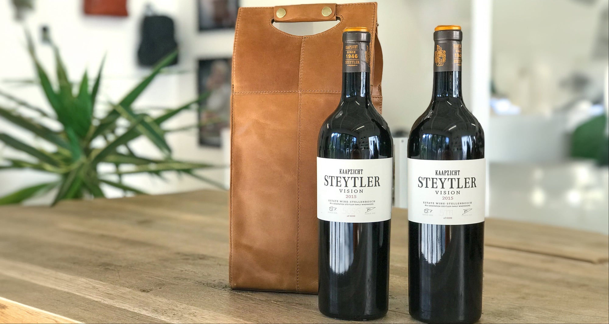 Two bottles of wine with a brown leather wine carrier on a wooden table.