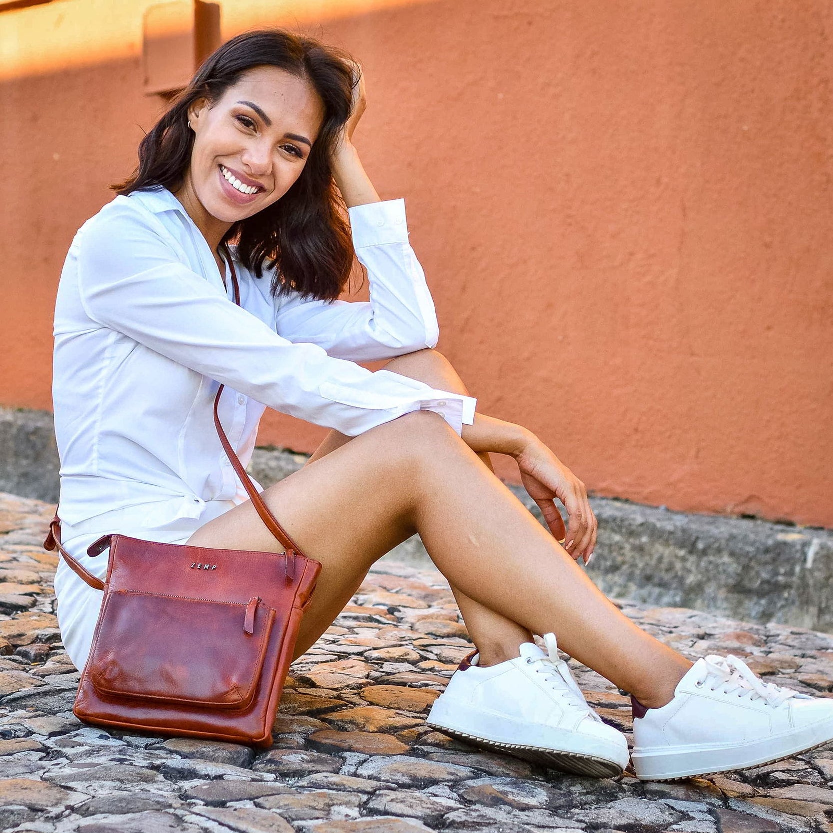 Woman sitting on a stone bench with a brown leather bag, wearing a white shirt and white sneakers.