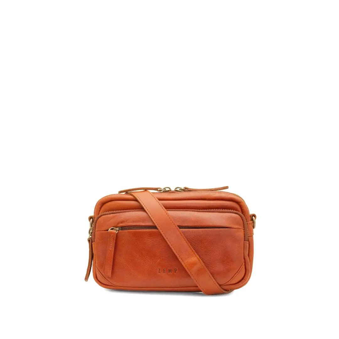 A warm chestnut leather crossbody bag with a matching leather shoulder strap, gold-tone hardware, and a front zippered pocket, shown on a plain white background.