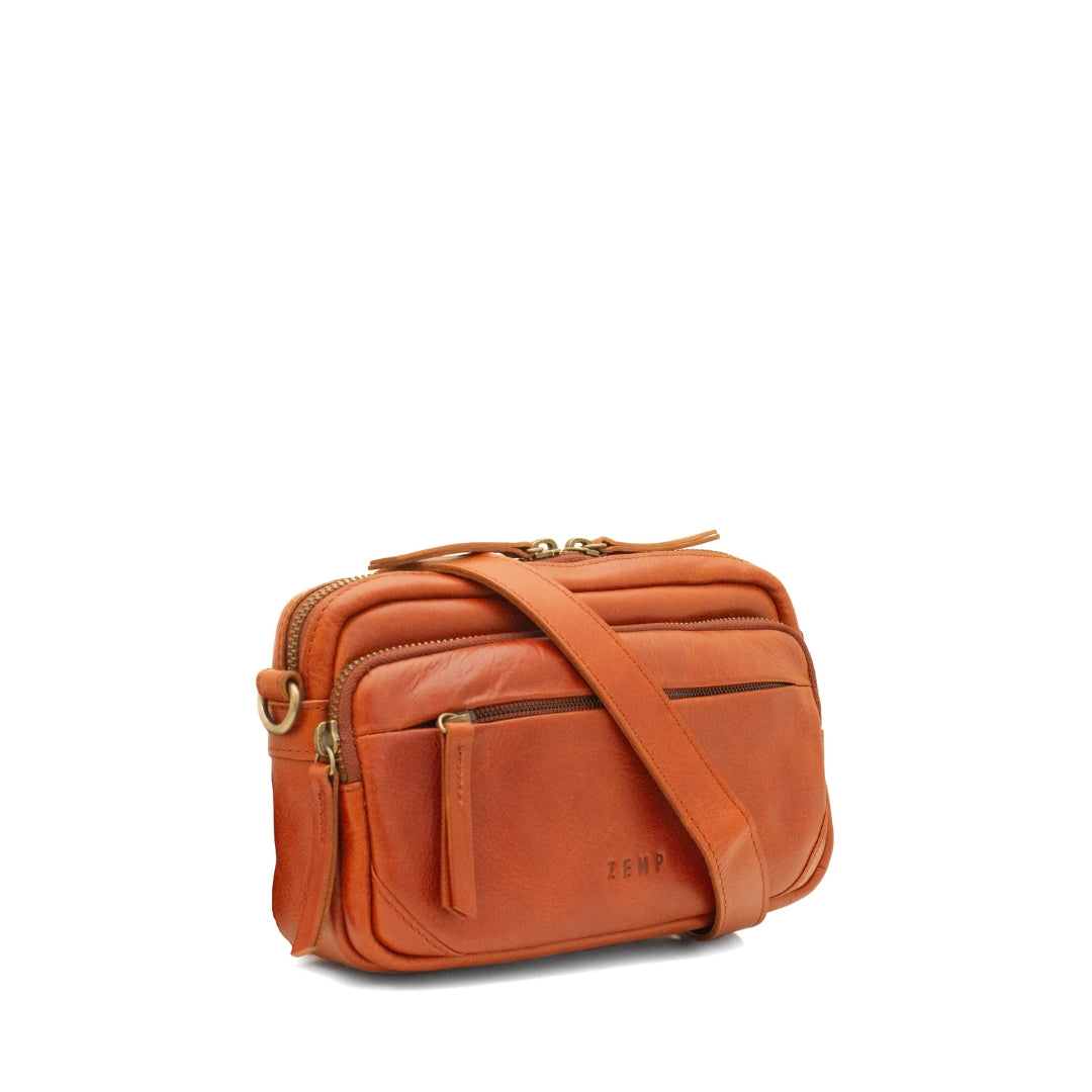 A warm chestnut leather crossbody bag with a matching leather shoulder strap, gold-tone hardware, and a front zippered pocket, shown on a plain white background.