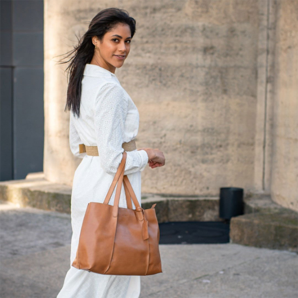 Woman in a white outfit holding a brown leather bag against a stone building background